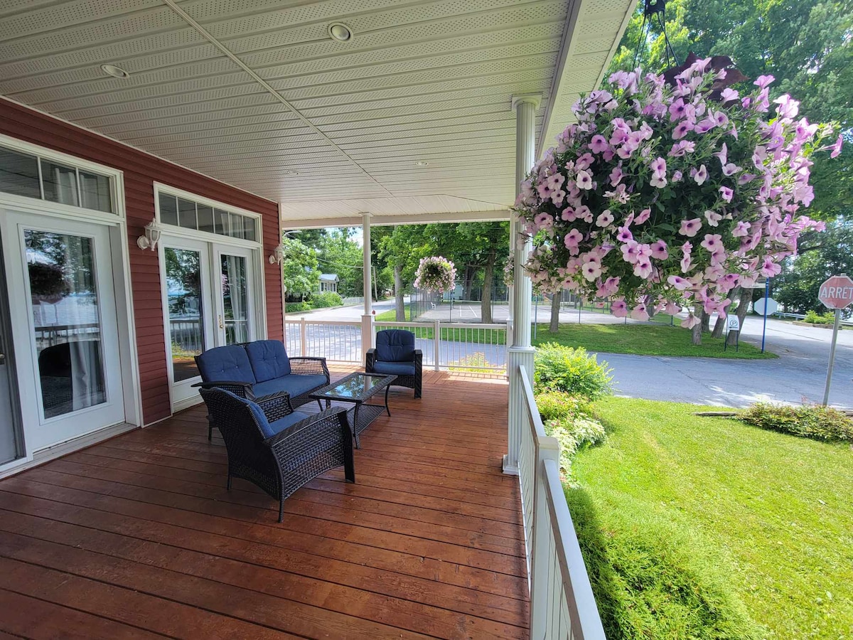A covered porch features a seating area with two dark wicker chairs and a glass-topped table. Hanging flower baskets filled with pink blooms add vibrancy to the space. Greenery surrounds the area, creating a calm, inviting atmosphere.