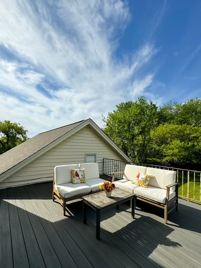 A roof deck is shown with two white cushioned chairs and a small table, adorned with a floral arrangement. The backdrop features a blue sky with scattered clouds and mature trees, providing a tranquil outdoor space.