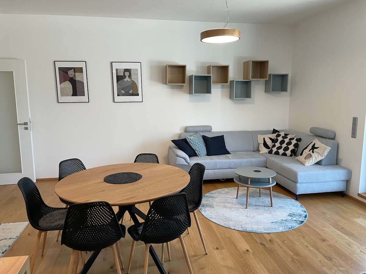 A contemporary living area features a light gray sectional sofa adorned with patterned cushions. A round wooden dining table is surrounded by six black chairs. Wall art and floating shelves add character, while a small round coffee table sits on a blue and white rug.