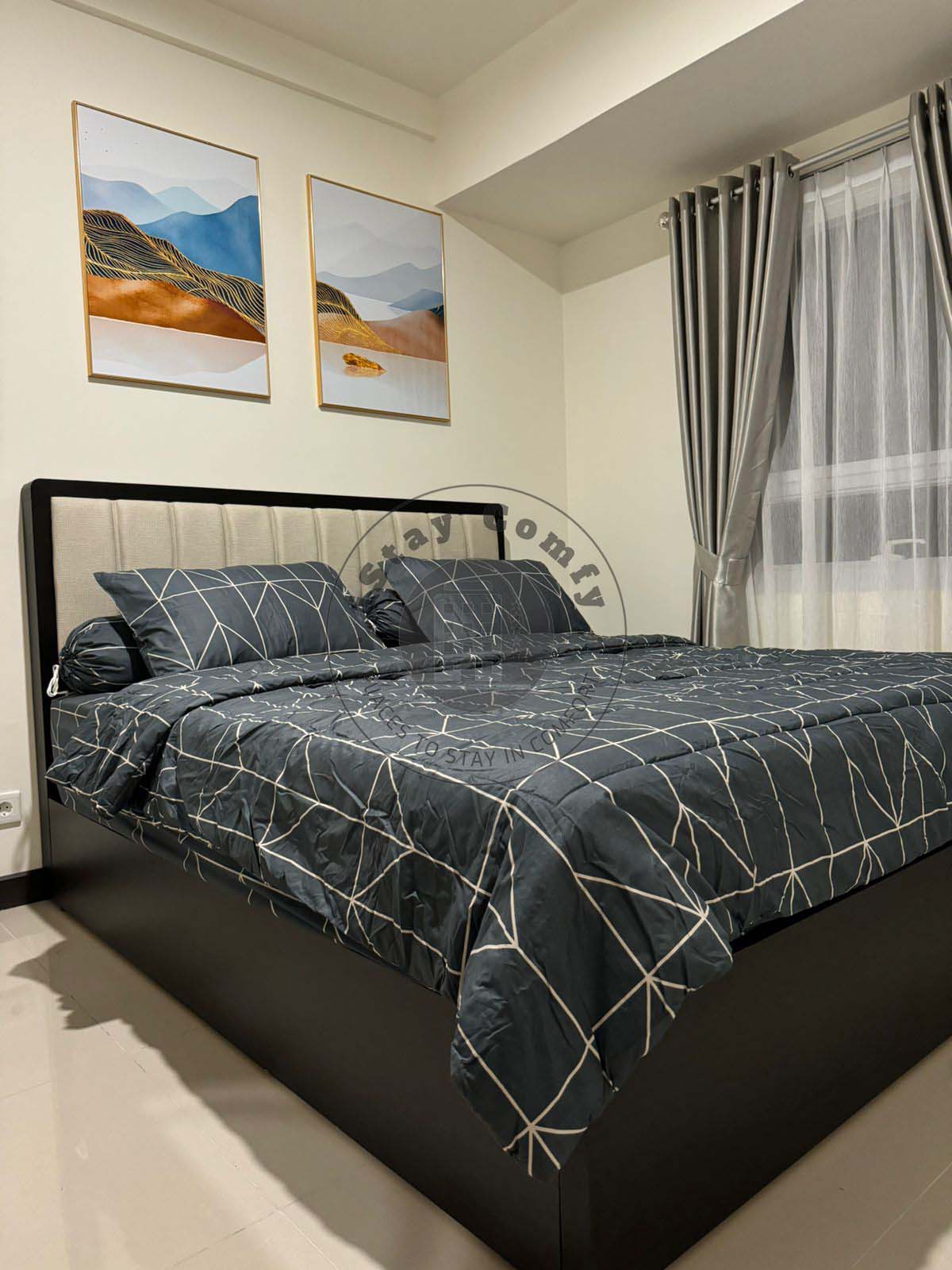 A spacious bed is adorned with a dark patterned bedspread and matching cushions. Above the bed, two framed artworks depict abstract landscapes. Sheer curtains allow natural light to enter, complementing the neutral-toned walls and sleek flooring.