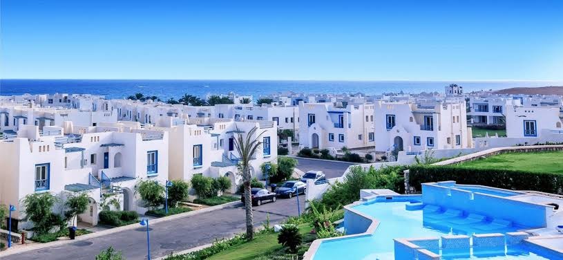 An expansive view captures the serene landscape, featuring a collection of white buildings arranged harmoniously among green palm trees. A shimmering infinity pool reflects the clear blue sky, with the coastline visible in the distance, offering a sense of tranquility.