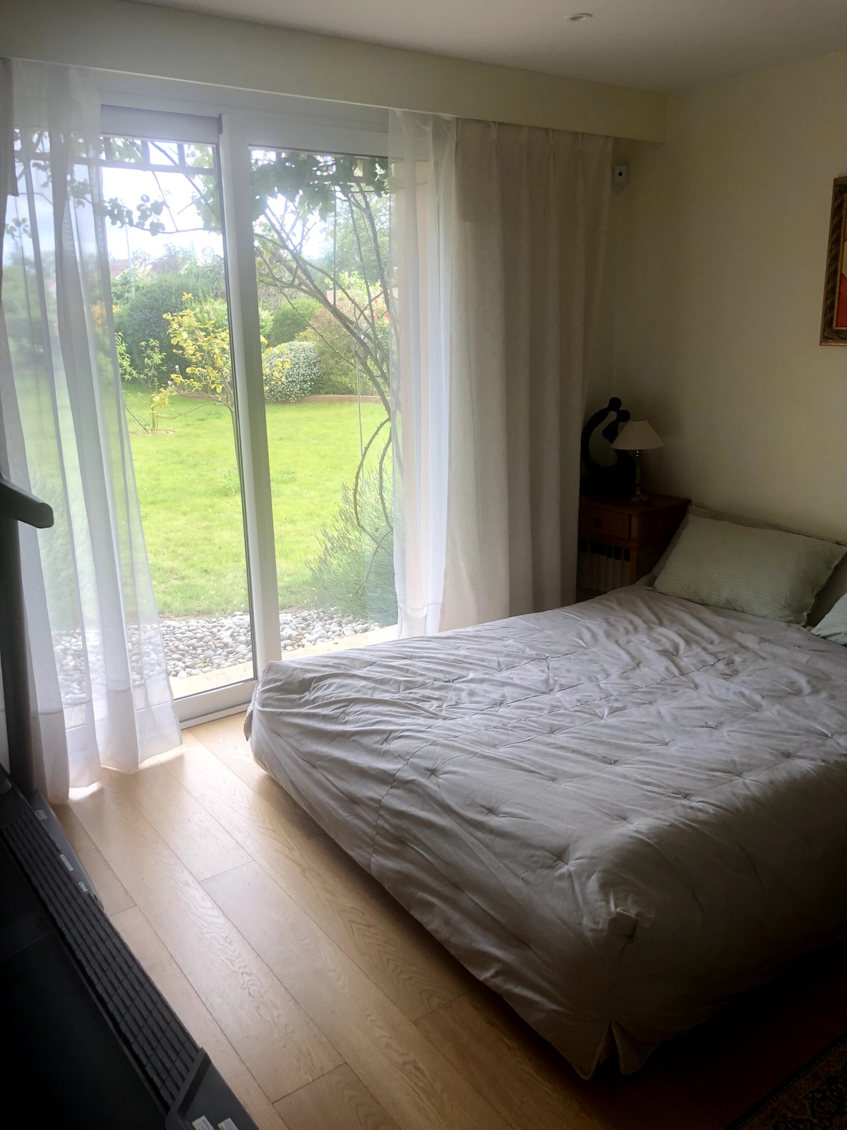 A spacious sleeping area features a light-colored bedspread atop a neatly made bed. Sheer curtains filter natural light from sliding glass doors that open to a grassy outdoor space, which is adorned with greenery and trees.