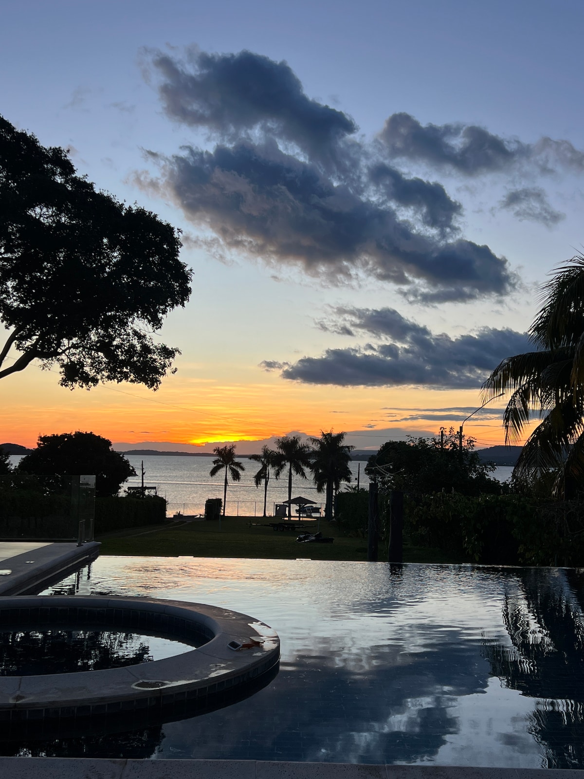 A serene view of the lake at sunset is captured, with the sky displaying soft hues of orange and purple. An infinity pool reflects the scenery, while palm trees line the waterfront. A hot tub is visible in the foreground, inviting relaxation.