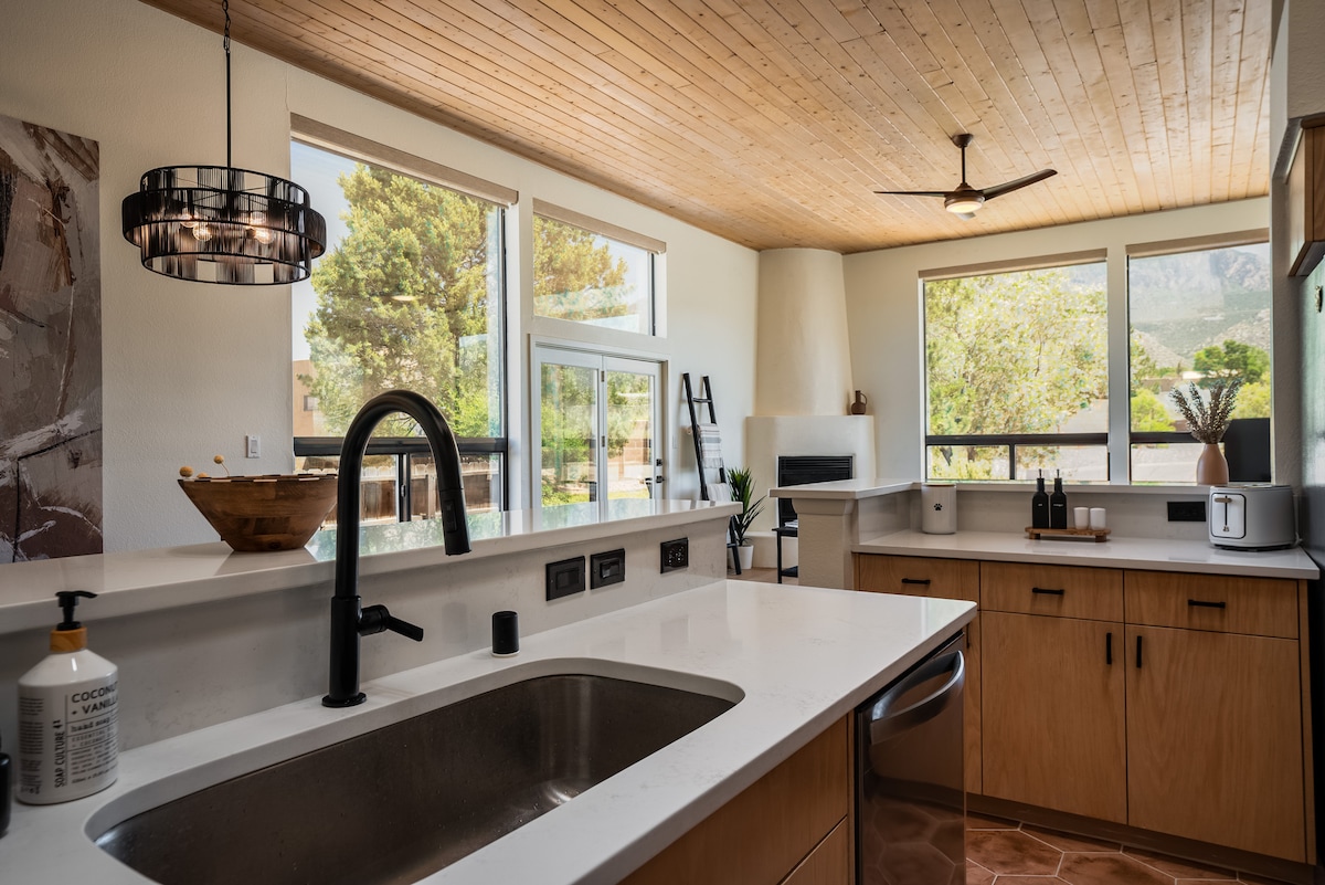 The kitchen features modern wooden cabinetry and a large island with a deep sink. Natural light enters through expansive windows, showcasing the serene outdoor landscape. A stylish pendant light hangs over the island, and stainless steel appliances are arranged for convenience.