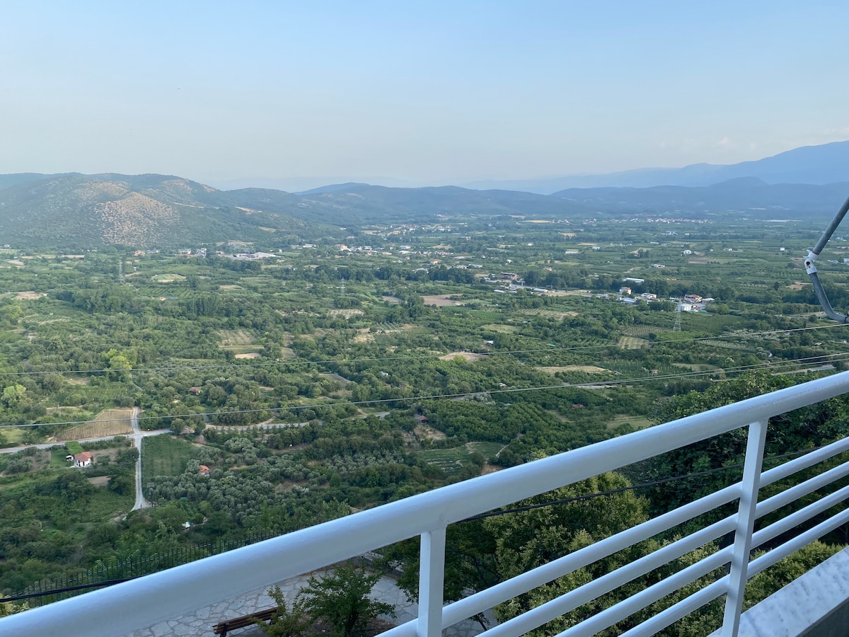 A panoramic view of lush green valleys and distant mountains is captured from the balcony. The expansive landscape features a patchwork of fields and orchards, with structures visible in the distance under a clear blue sky.
