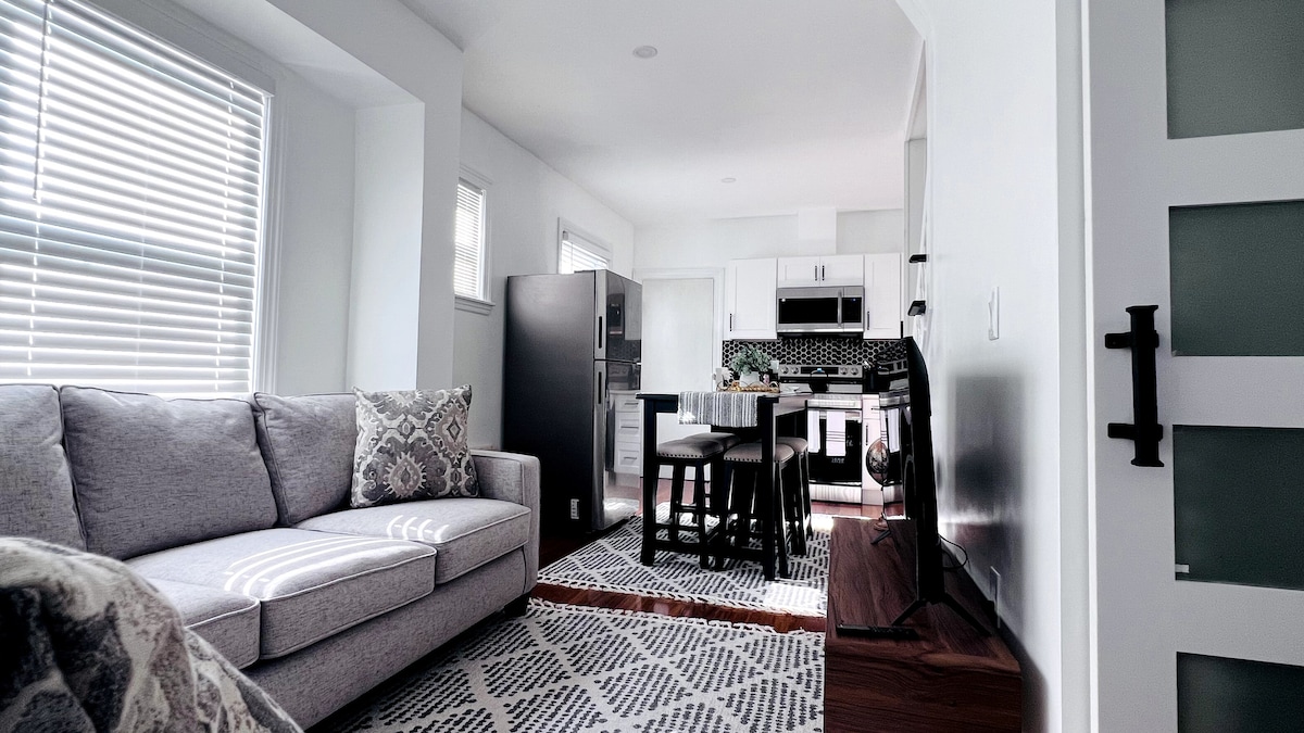The open living area features a light gray sofa adorned with decorative pillows. A dining table with high-backed seating is visible, alongside modern appliances in the fully equipped kitchen. Natural light enters through multiple windows, illuminating the harmonious space.