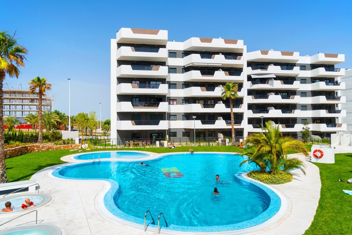 A modern apartment complex is showcased, featuring a curvilinear pool surrounded by a well-manicured landscape. Sun loungers line the edge of the pool, where guests can be seen enjoying the water. Palm trees are visible in the background against a clear blue sky.