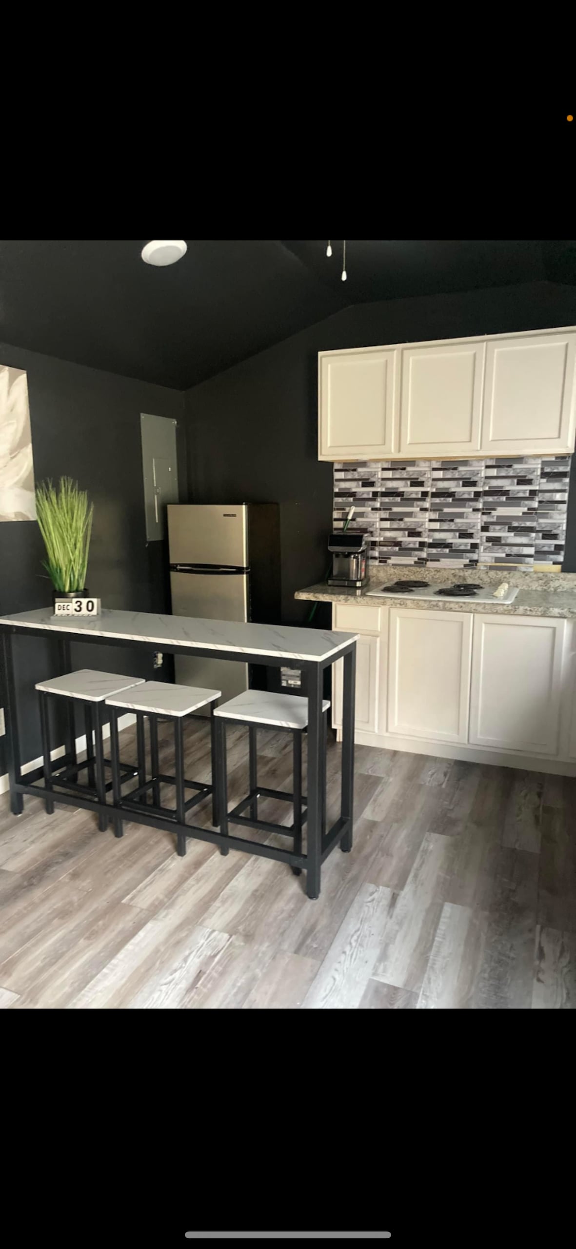 The kitchen features a modern design with a black accent wall and light cabinetry. There is a small dining table with three stools, and an updated silver refrigerator is visible. A countertop with a stone backsplash complements the functional appliances in the space.