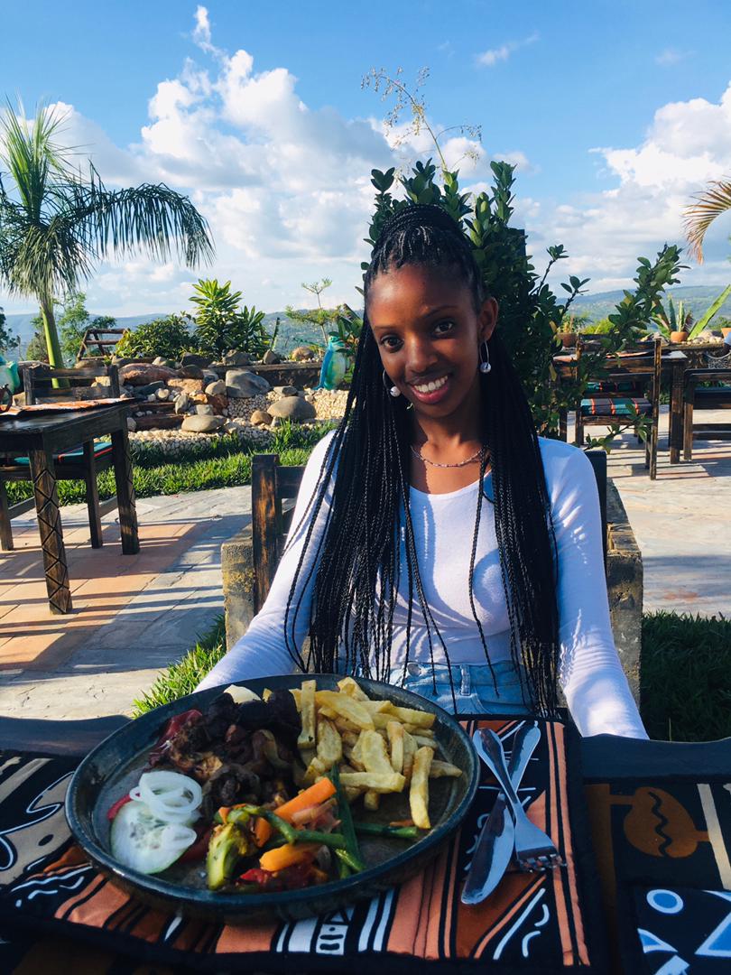 A dining table is set outdoors, featuring a plate of assorted food including vegetables and fries. Lush greenery and decorative plants are visible in the background, alongside wooden dining furniture and a clear blue sky with scattered clouds.