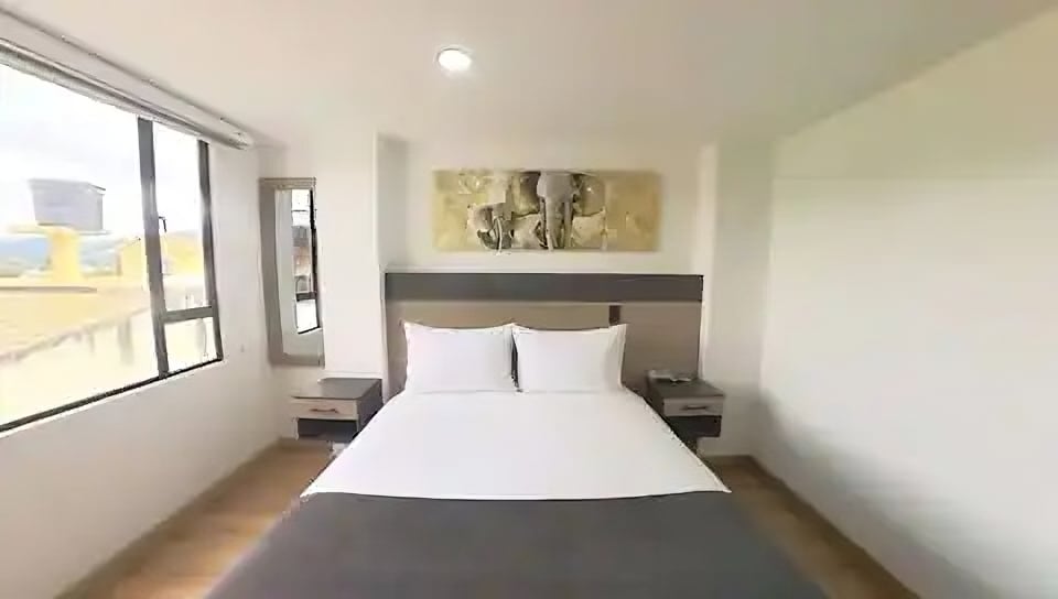 A modern bedroom features a spacious bed with crisp white linens and a decorative wall art piece above the headboard. Large windows allow natural light to fill the room, while wooden flooring adds warmth to the space.
