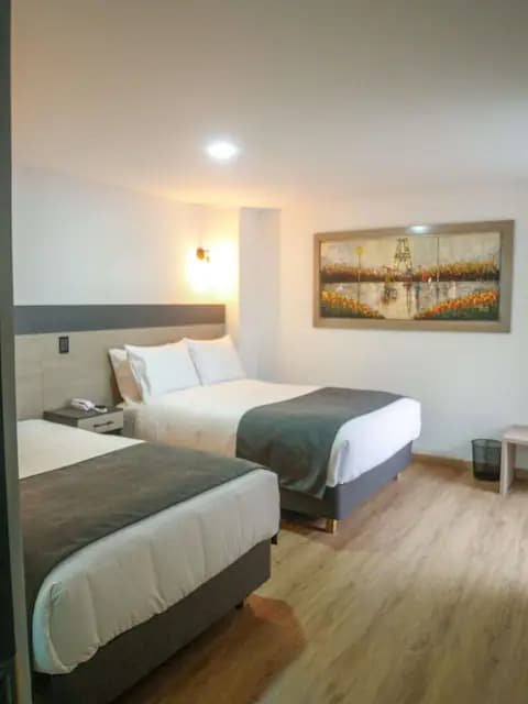 A modern guest room features two spacious beds with crisp white linens and grey bed throws. A piece of artwork depicting a serene landscape adorns one wall. Soft lighting is provided by sconces, and a small trash bin is placed discreetly in the corner.