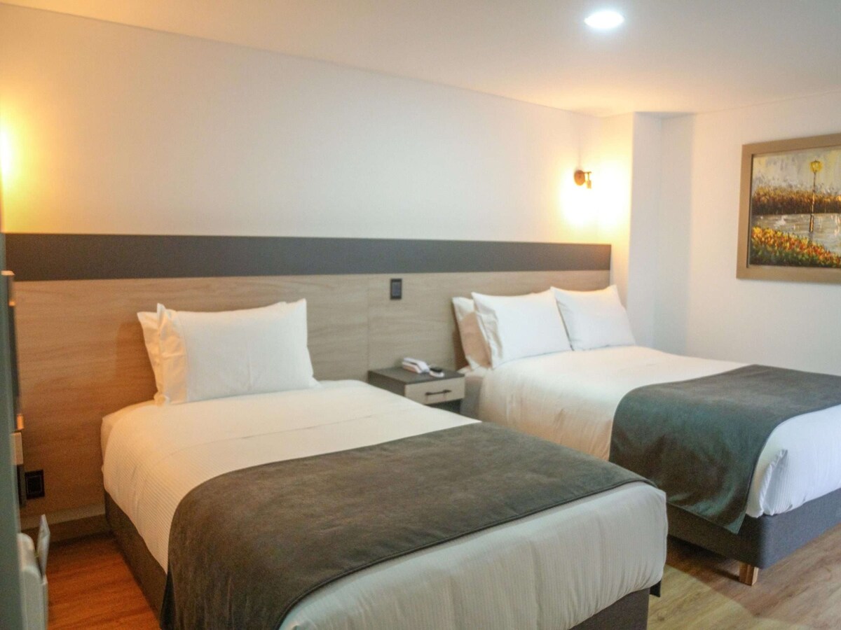 A well-appointed room features two spacious beds with neutral-toned linens. Soft lighting from wall sconces illuminates the space, while a small bedside table sits between the beds. A piece of artwork hangs on the wall, adding a touch of color to the modern decor.