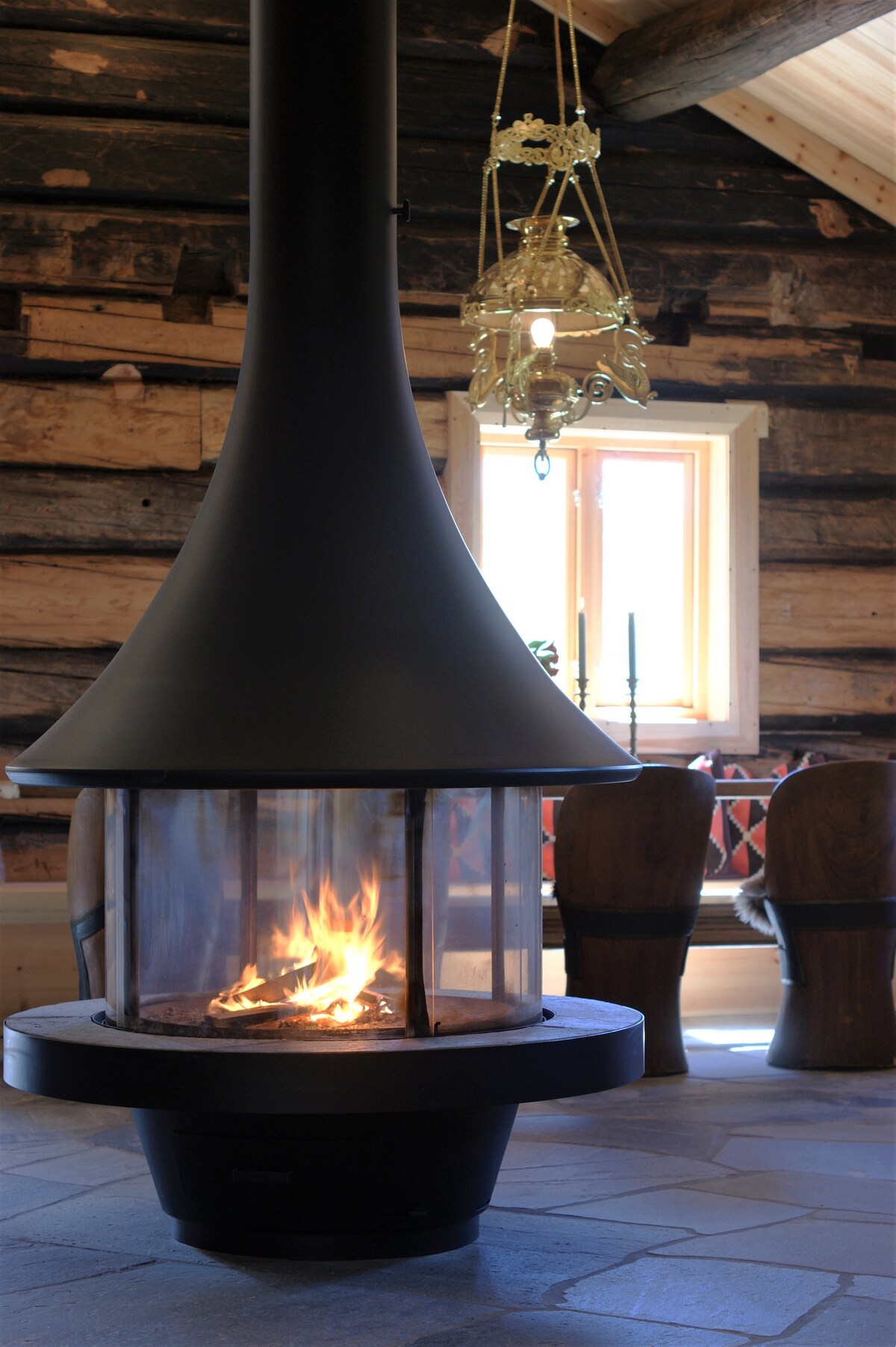 A modern wood stove features a sleek design with a glass enclosure, displaying a gentle flame. Warm light from an antique chandelier above complements the rustic wooden backdrop. A dining area with large wooden chairs is set in the background, enhancing the inviting atmosphere.