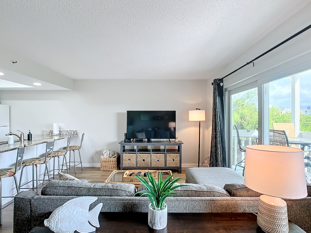 An inviting living area features a large sectional sofa and a flat-screen television mounted on the wall. Natural light fills the space through expansive glass doors that open to a balcony. A warm lamp and a decorative plant enhance the cozy ambiance.