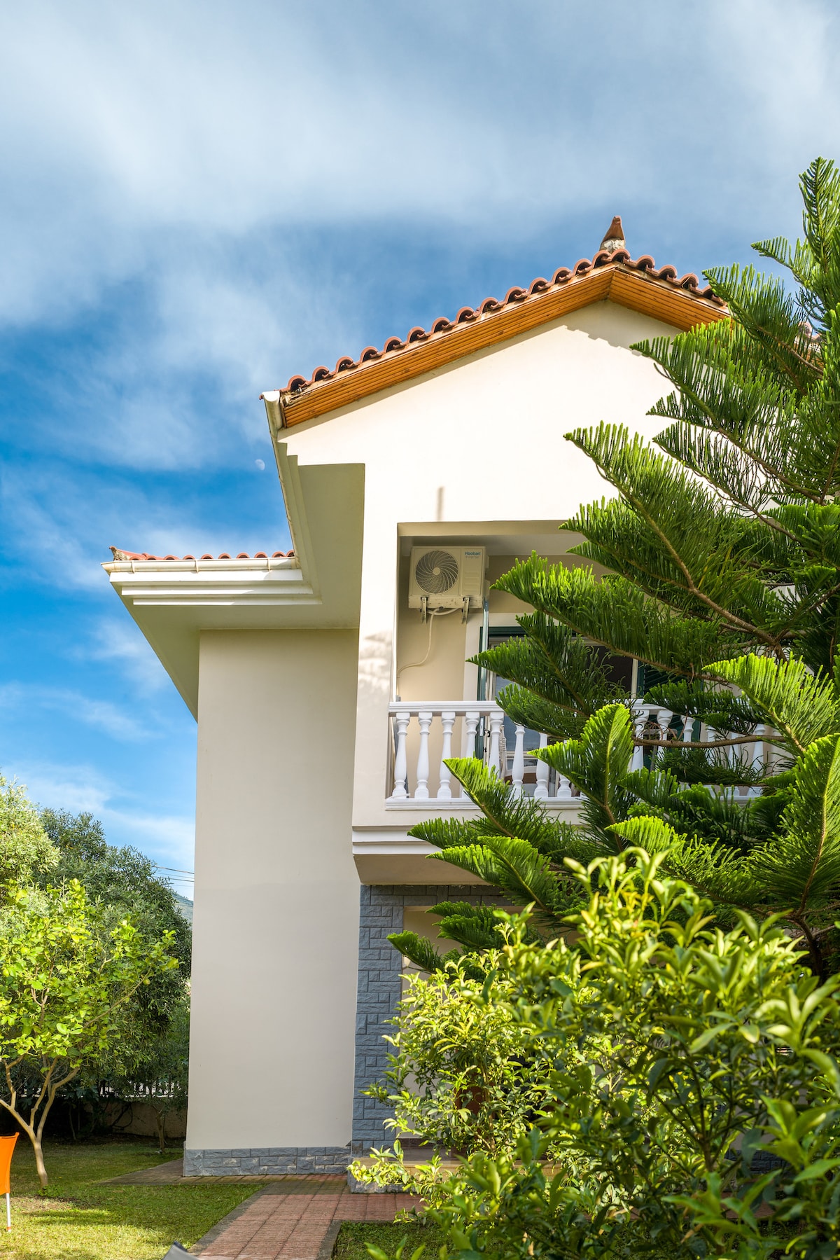 The exterior of the villa is shown, featuring a two-story design with a terracotta roof. Lush greenery surrounds the structure, including tall pine trees and well-kept shrubs. An air conditioning unit is mounted on the wall, providing comfort during warm weather.