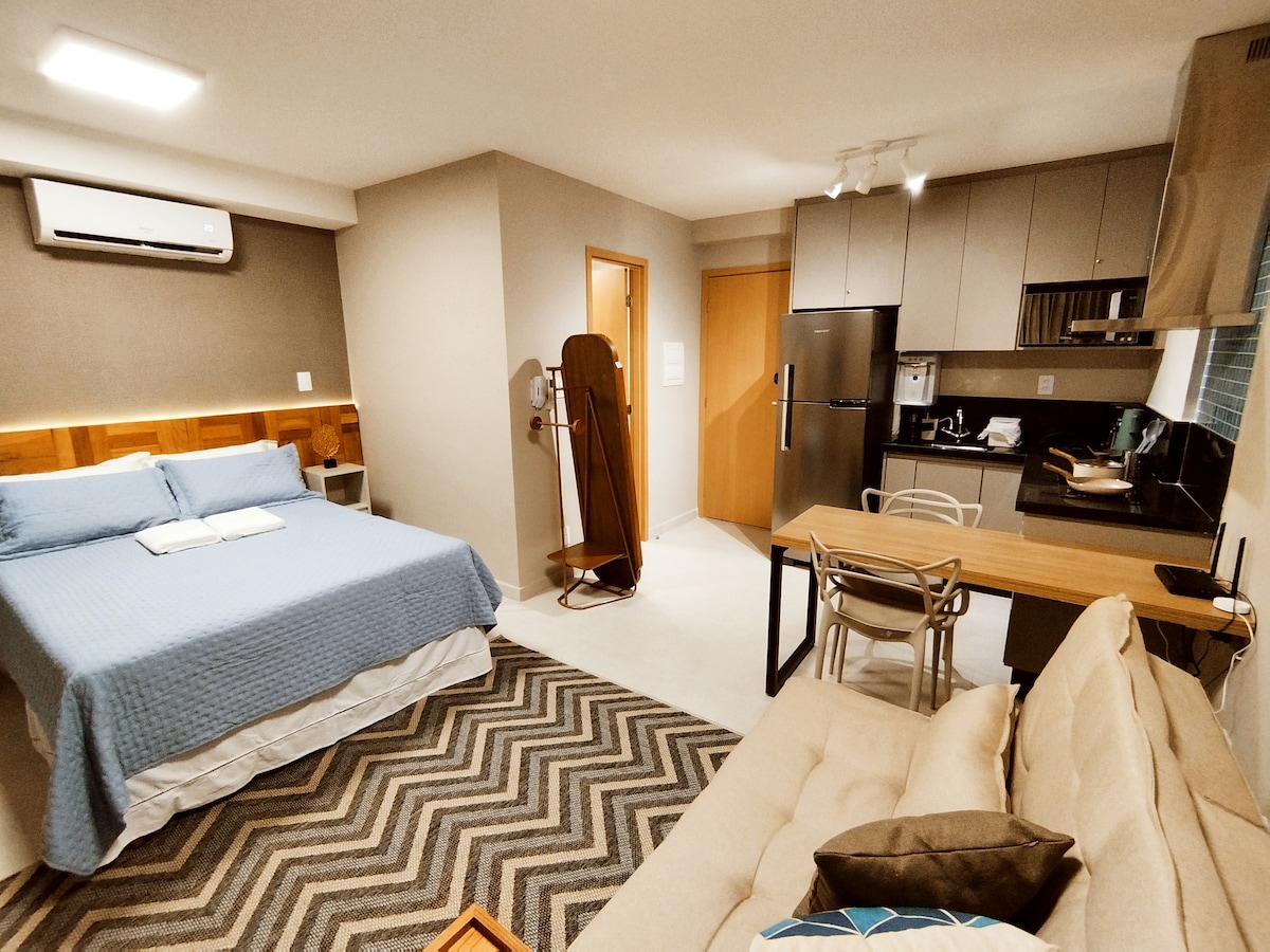 A cozy studio apartment features a spacious bed with a blue coverlet, complemented by a patterned rug. A compact kitchen is situated against one wall, next to a dining table and chairs. Natural light enters from a nearby window, enhancing the welcoming ambiance.