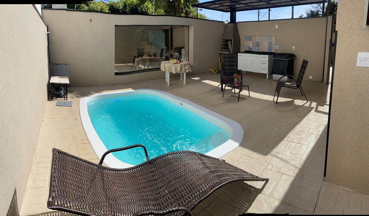 A private outdoor space features a refreshing pool surrounded by a sun deck. Comfortable lounge chairs are positioned nearby, and a shaded area includes outdoor furniture and a grill. Large windows offer a view of the pool, enhancing the inviting atmosphere.