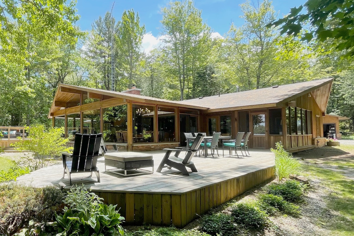 A spacious wooden deck surrounds the lakefront home, featuring a mix of cozy seating, including lounge chairs and dining tables. Large windows allow natural light to fill the interior while offering glimpses of the surrounding greenery and mature trees.