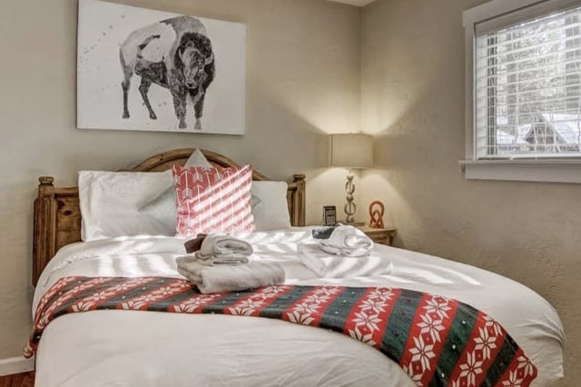 A cozy bedroom features a large bed adorned with crisp white linens and a festive red and green blanket. A buffalo wall art piece adds character, while soft light from a bedside lamp illuminates the room. Fresh towels are neatly arranged on the bed.