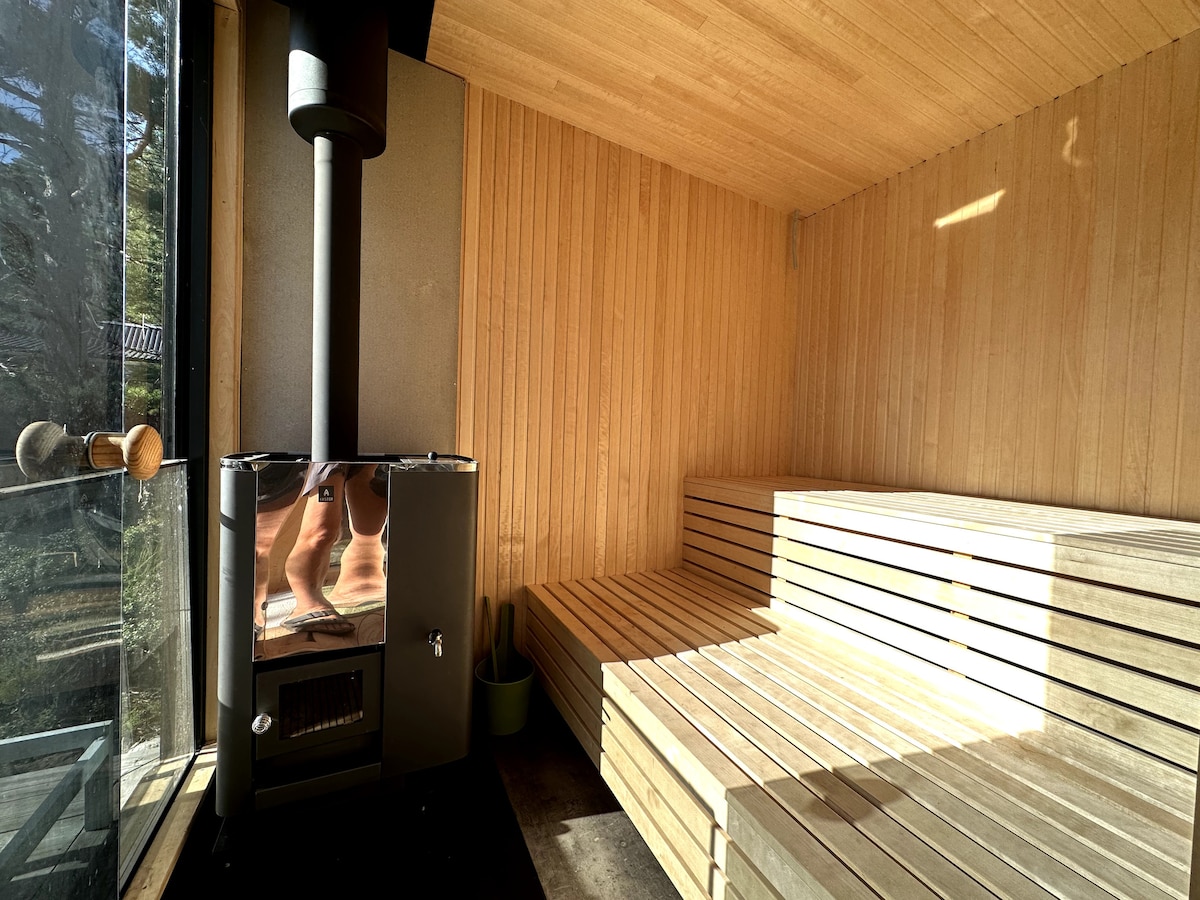 A wooden sauna features a cozy interior with benches designed for relaxation. A stove is positioned in the corner, providing warmth. Large windows allow natural light to enter, offering a view of the surrounding nature.
