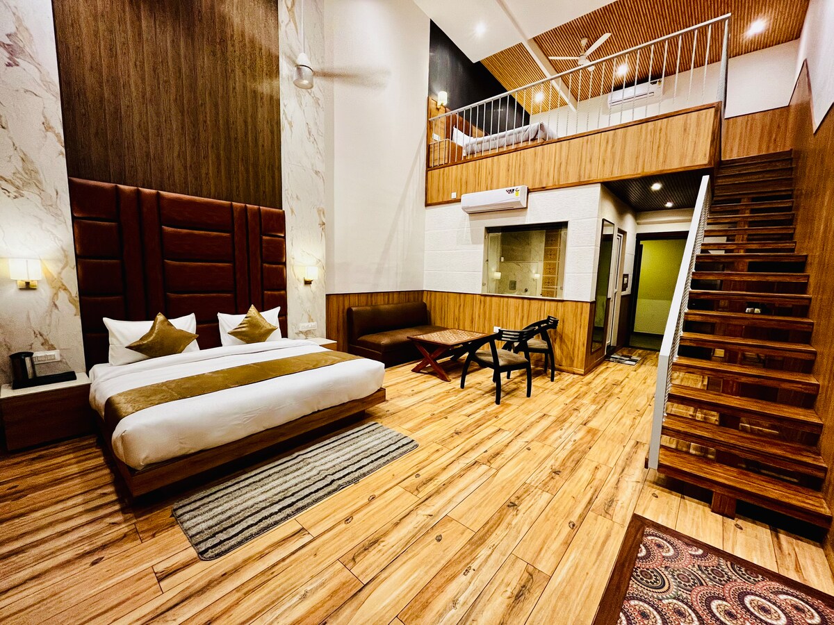 A spacious room features two double beds with neutral-toned bedding. Wooden flooring complements the contemporary design, while a seating area offers a low table and two chairs. An open staircase leads to a loft area above, and a glass-enclosed bathroom area is visible.