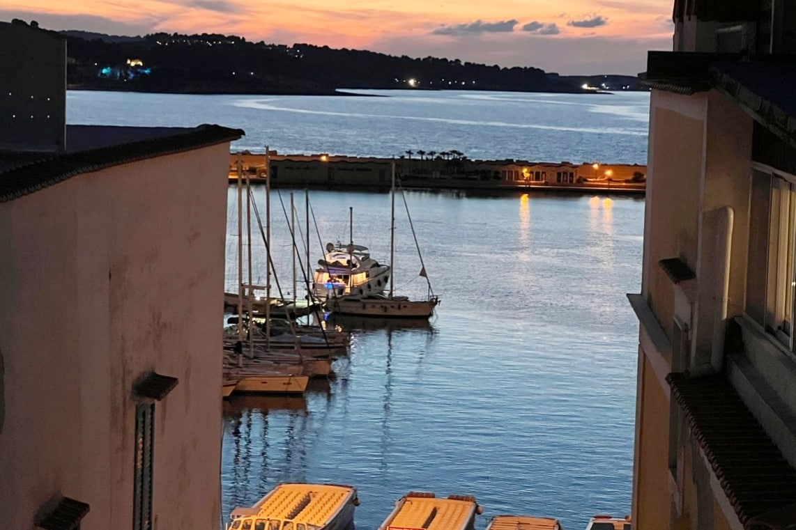A serene view captures the marina at dusk, with several sailing boats gently anchored. The water reflects warm hues from the sunset, while softly illuminated buildings line the shore, adding to the peaceful ambiance of the scene.