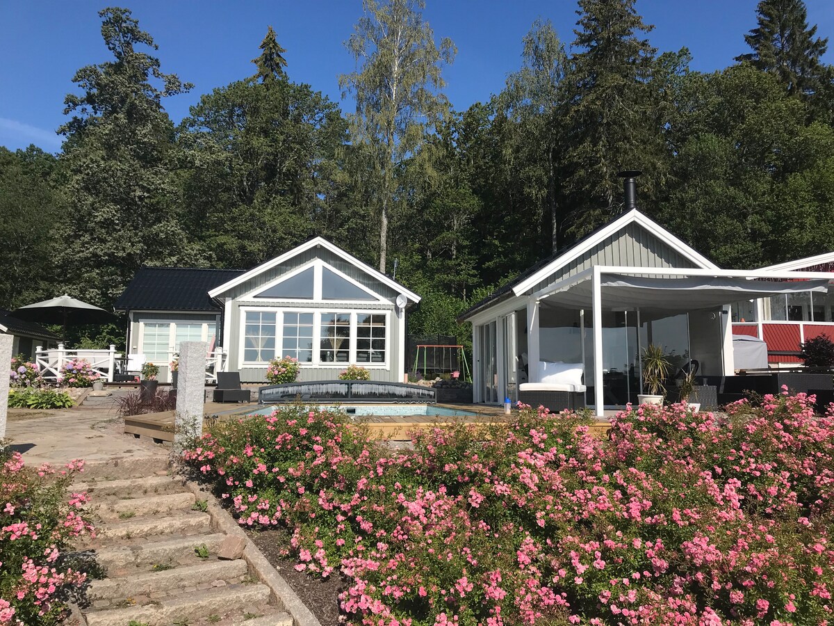 A charming exterior view of a family-friendly home is framed by blooming pink flowers. The structure features large windows, allowing natural light to enter, while outdoor seating is visible on the patio. Lush greenery provides a scenic backdrop to the inviting space.