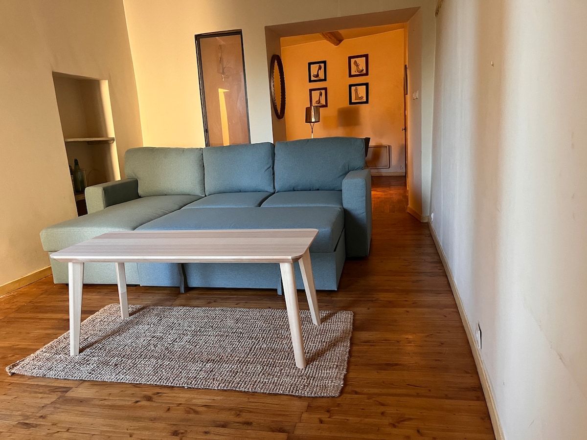 A spacious living area features a light blue sectional sofa and a wooden coffee table placed on a woven rug. Warm wooden flooring complements the room, which is illuminated by natural light. An adjoining area reveals framed wall art and a lamp in the background.