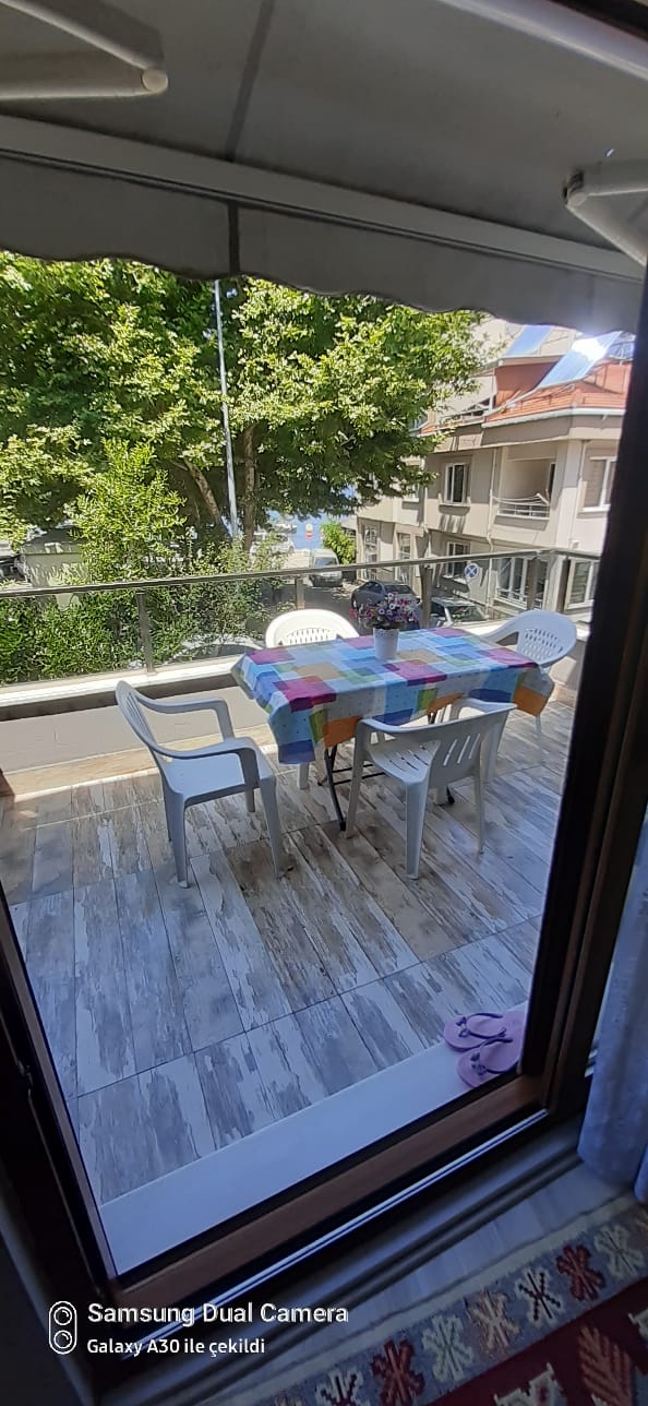 A spacious balcony features a table set with colorful tableware, surrounded by several chairs. Lush greenery is visible beyond a glass railing, with a glimpse of the sea in the distance, enhancing the outdoor dining experience.