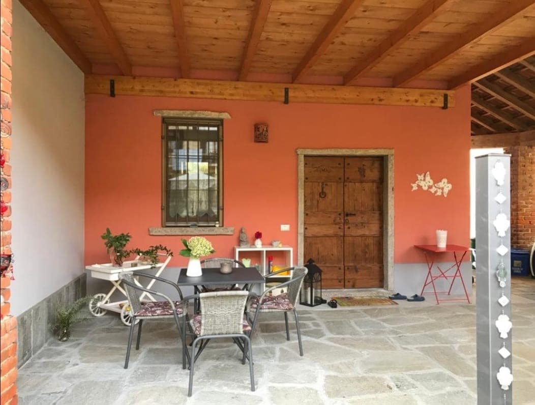 A covered outdoor sitting area features a stone floor and wooden beams overhead. A rectangular table with black chairs is set for dining, accompanied by planters. The charming red wall is accentuated by a rustic wooden door and window, inviting natural light into the space.