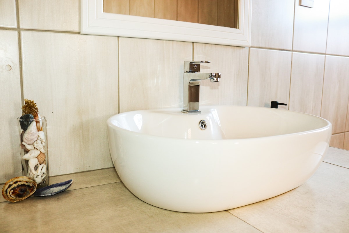 A modern bathroom sink with a sleek, contemporary design is displayed. The oval bowl is set upon a smooth countertop, accompanied by a polished faucet. A small glass container filled with shells adds a decorative touch nearby.