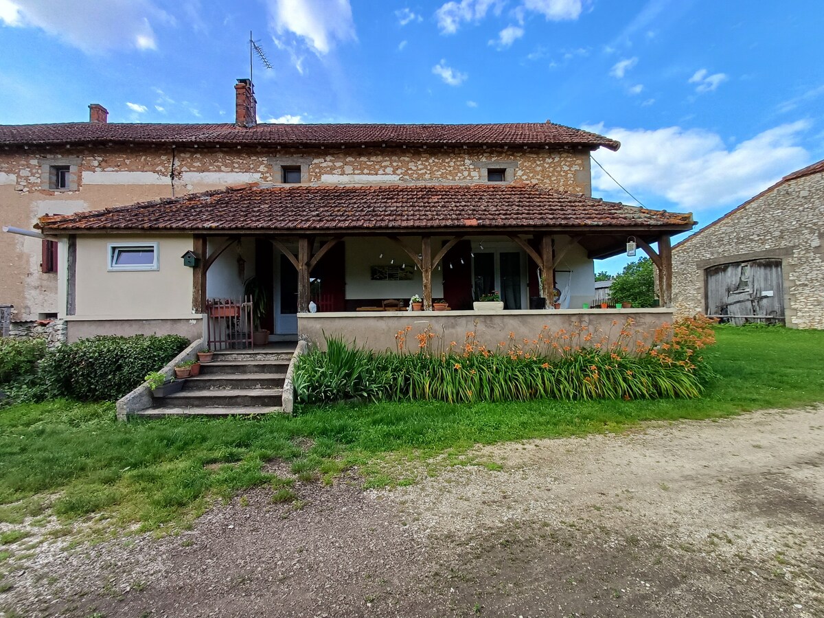 The exterior of the cottage showcases a covered terrace that features wooden beams and a sloped tile roof. A pathway leads to the entrance, flanked by vibrant orange flowers and green grass. The structure combines rustic stone and plaster finishes under a partly cloudy sky.