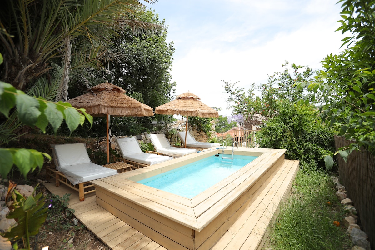 A shared swimming pool is surrounded by wooden decking, featuring steps for easy entry. Comfortable lounge chairs are placed beneath thatched umbrellas, offering shade. Lush greenery encircles the pool area, enhancing the tranquil atmosphere.