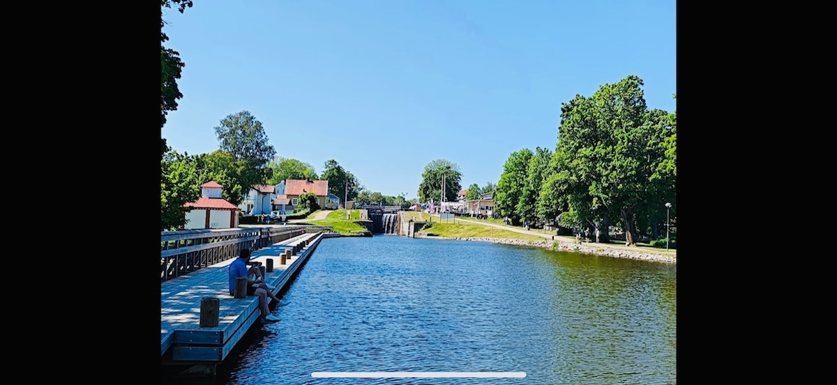 A calm body of water is framed by lush green trees and a clear blue sky. On the left, a wooden dock juts into the water, while a historical lock system can be seen in the distance. Quaint buildings peek through the foliage, highlighting the serene setting.