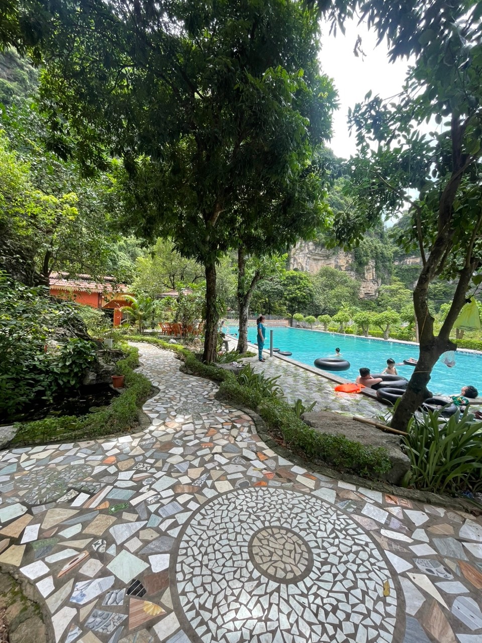 A winding stone pathway leads through lush greenery towards a turquoise pool, where guests are seen relaxing on floating inflatables. Towering trees provide shade, and a rocky landscape is visible in the background, contributing to a serene outdoor atmosphere.
