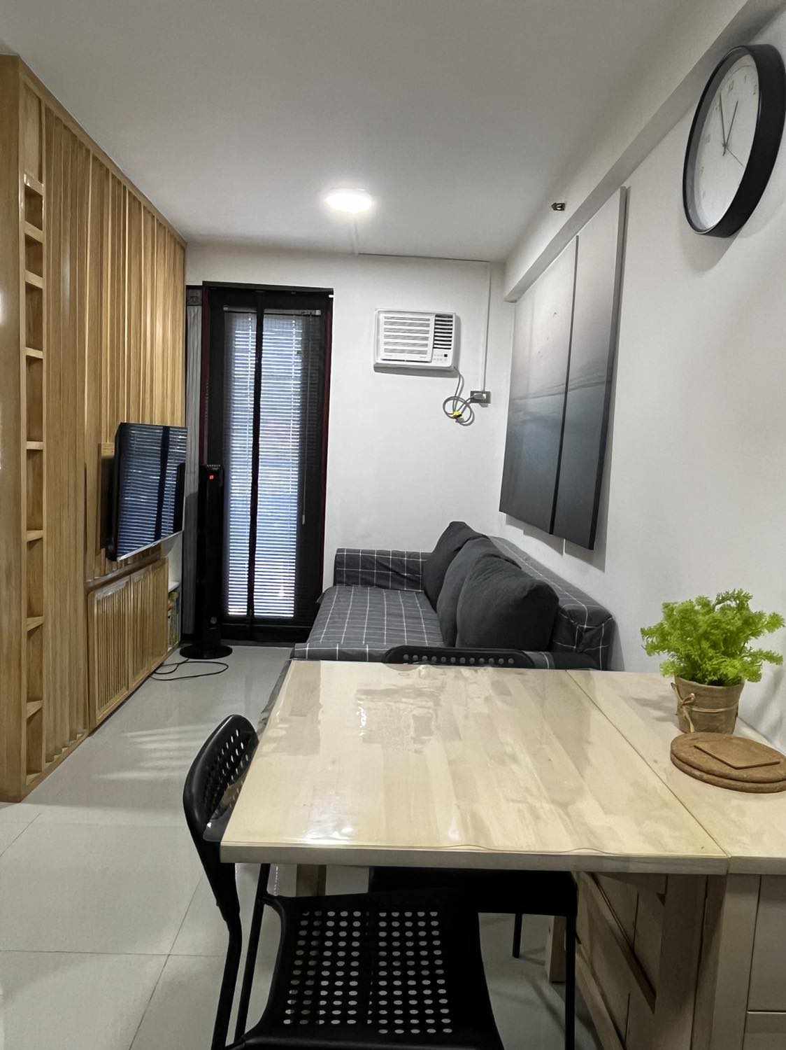 A living area features a comfortable gray sofa positioned next to a modern TV unit. A small dining table with black chairs is visible, providing a space for dining or work. Large windows allow natural light while providing views of the outside.