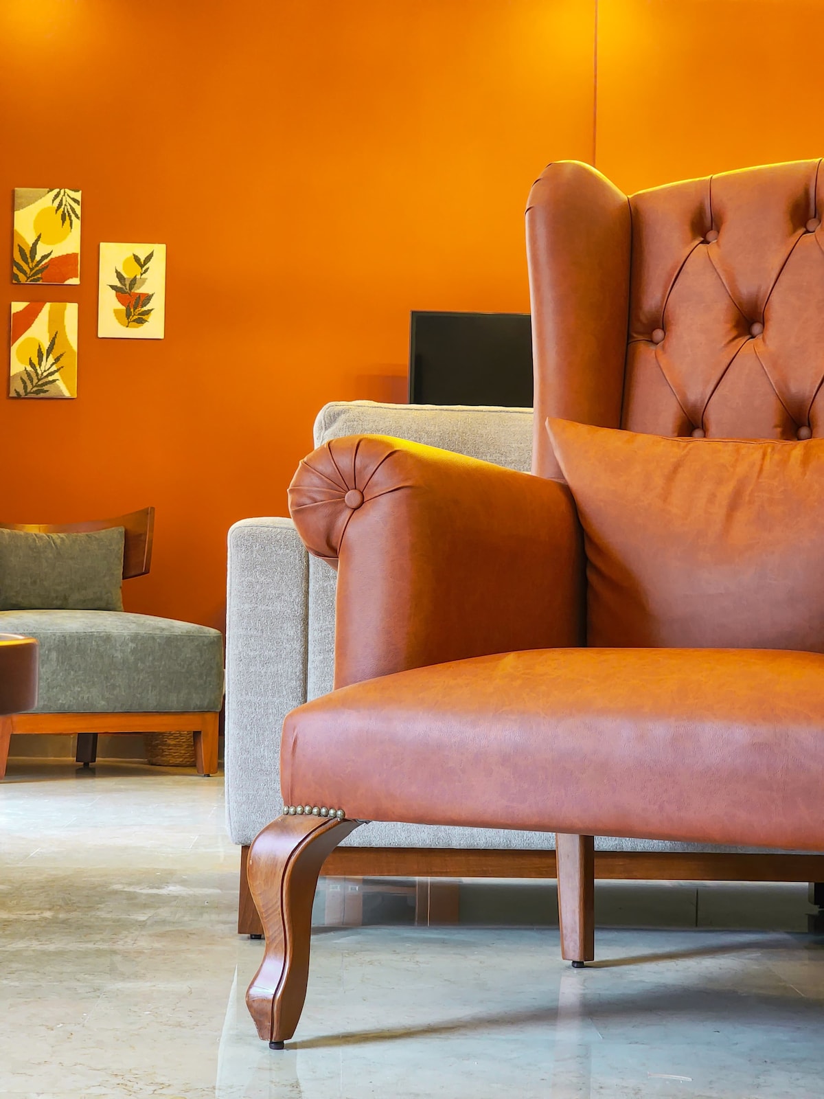 A brown leather armchair, featuring tufted detailing and elegant wooden legs, is positioned prominently in the living area. The warm orange wall adds vibrancy, while abstract wall art complements the cozy seating arrangement, which includes a light gray sofa in the background.
