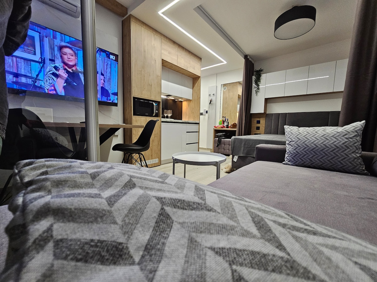 A modern open-space living area features a comfortable sectional sofa with patterned cushions and a circular table. A sleek kitchen area is visible in the background, with modern appliances. A television mounted on the wall displays a program, while soft ambient lighting enhances the space.