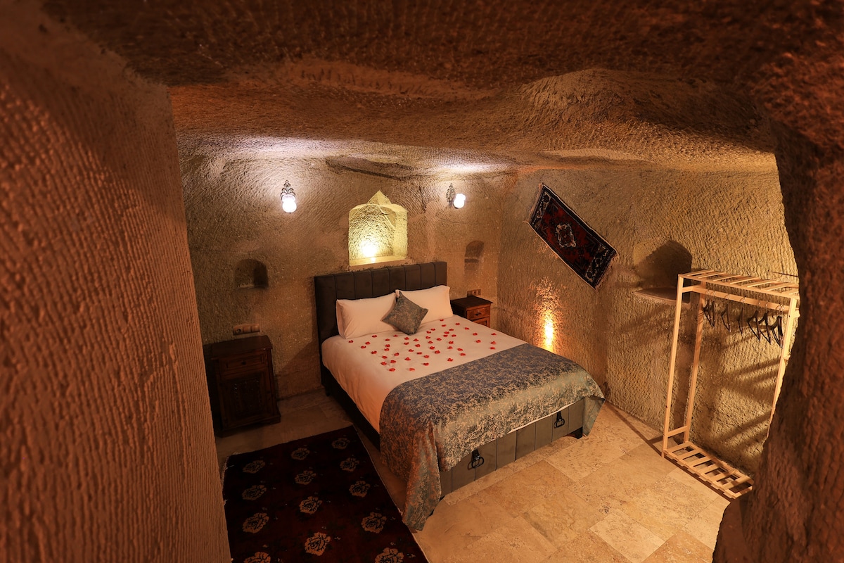A cave room features a spacious bed adorned with decorative pillows and a patterned bedspread. Soft lighting is provided by wall-mounted lanterns, with textured stone walls surrounding the space. A traditional rug enhances the floor, and a simple clothing rack is positioned in one corner.