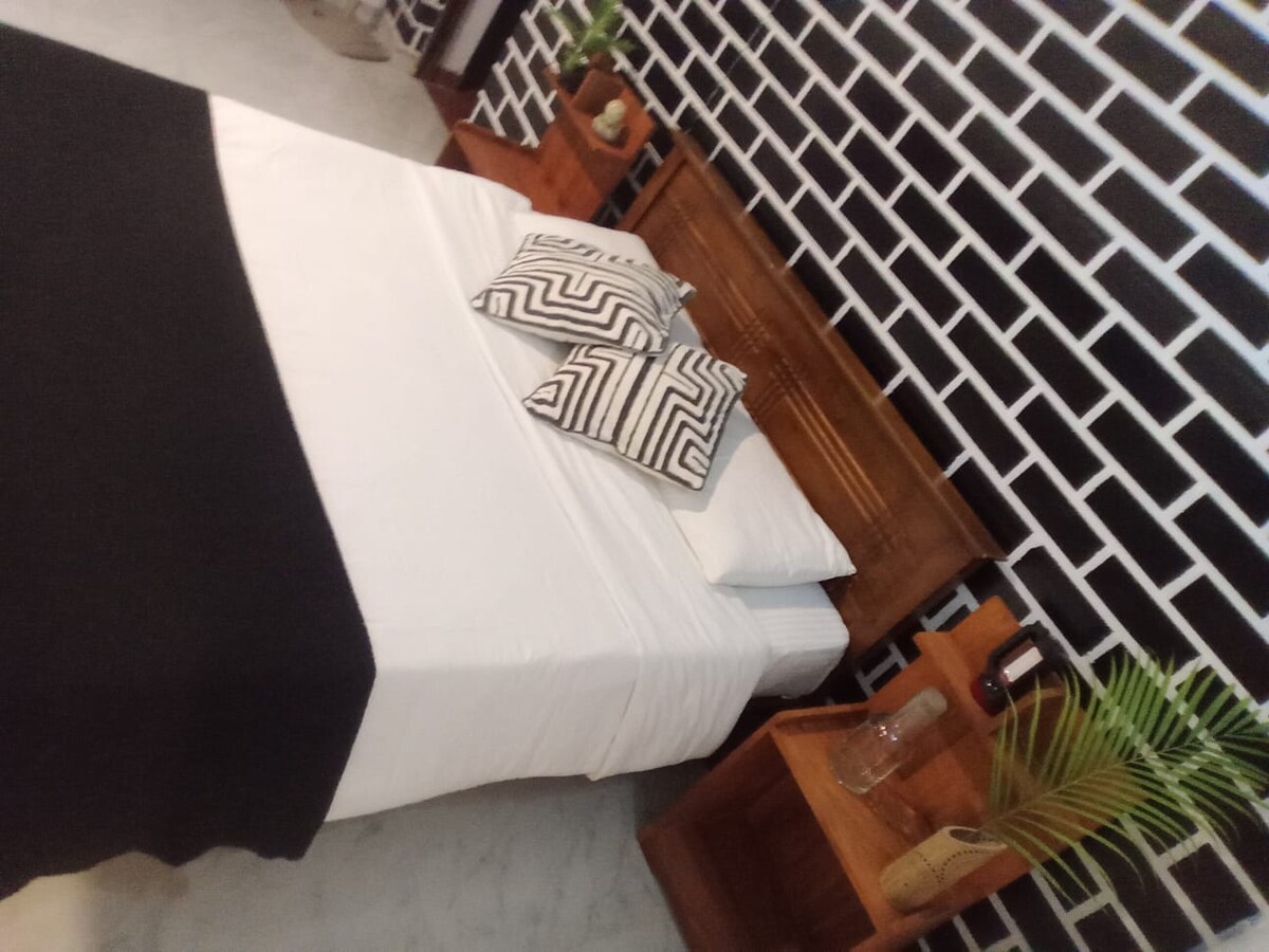 A minimalist bedroom is presented with a spacious bed dressed in white linens, complemented by contrasting black accents. Geometric-patterned pillows are placed atop the bed, with wooden nightstands featuring small plants and decorative items on each side. A brick-patterned wall adds texture to the space.