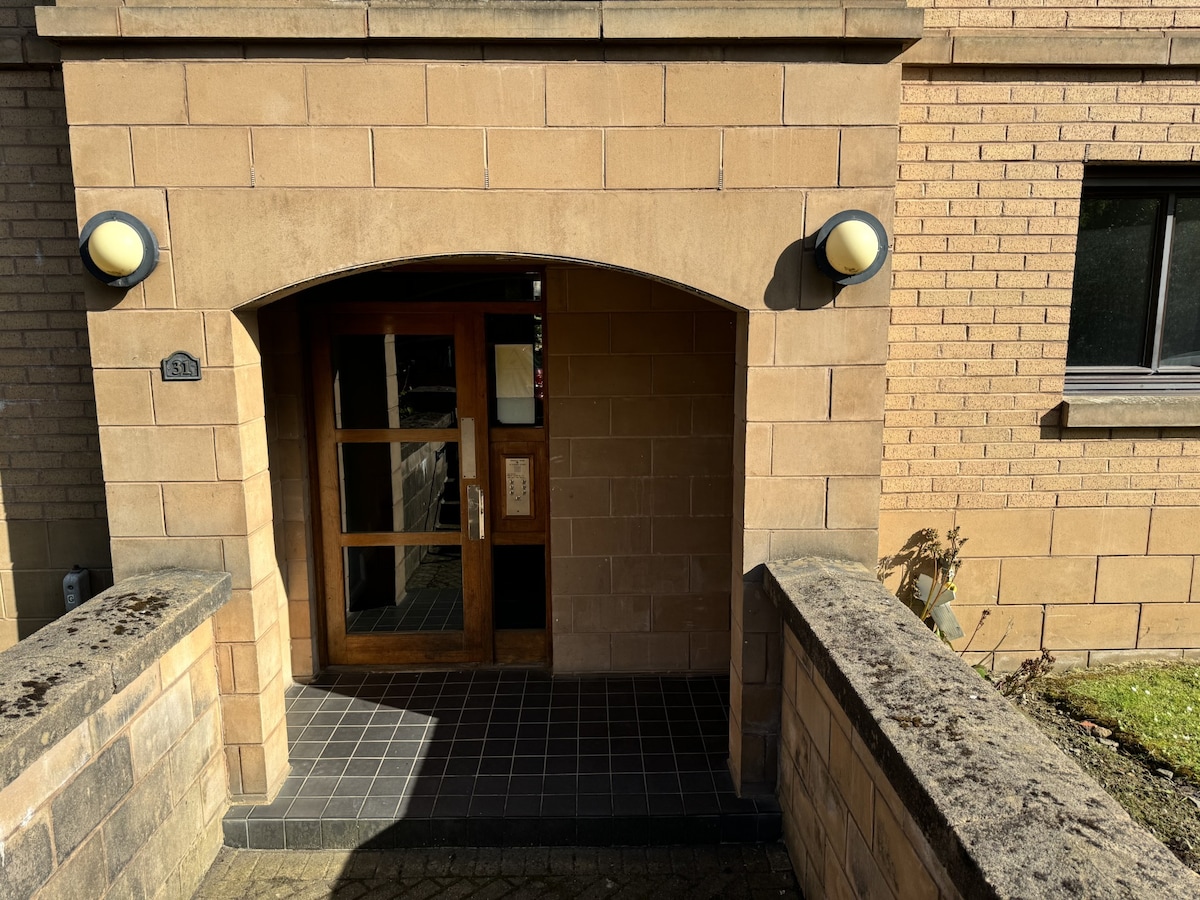 The entrance features a large wooden door framed by an archway, illuminated by two circular wall sconces. A set of stone steps leads up to the door from the paved pathway, providing an inviting entry point to the apartment.