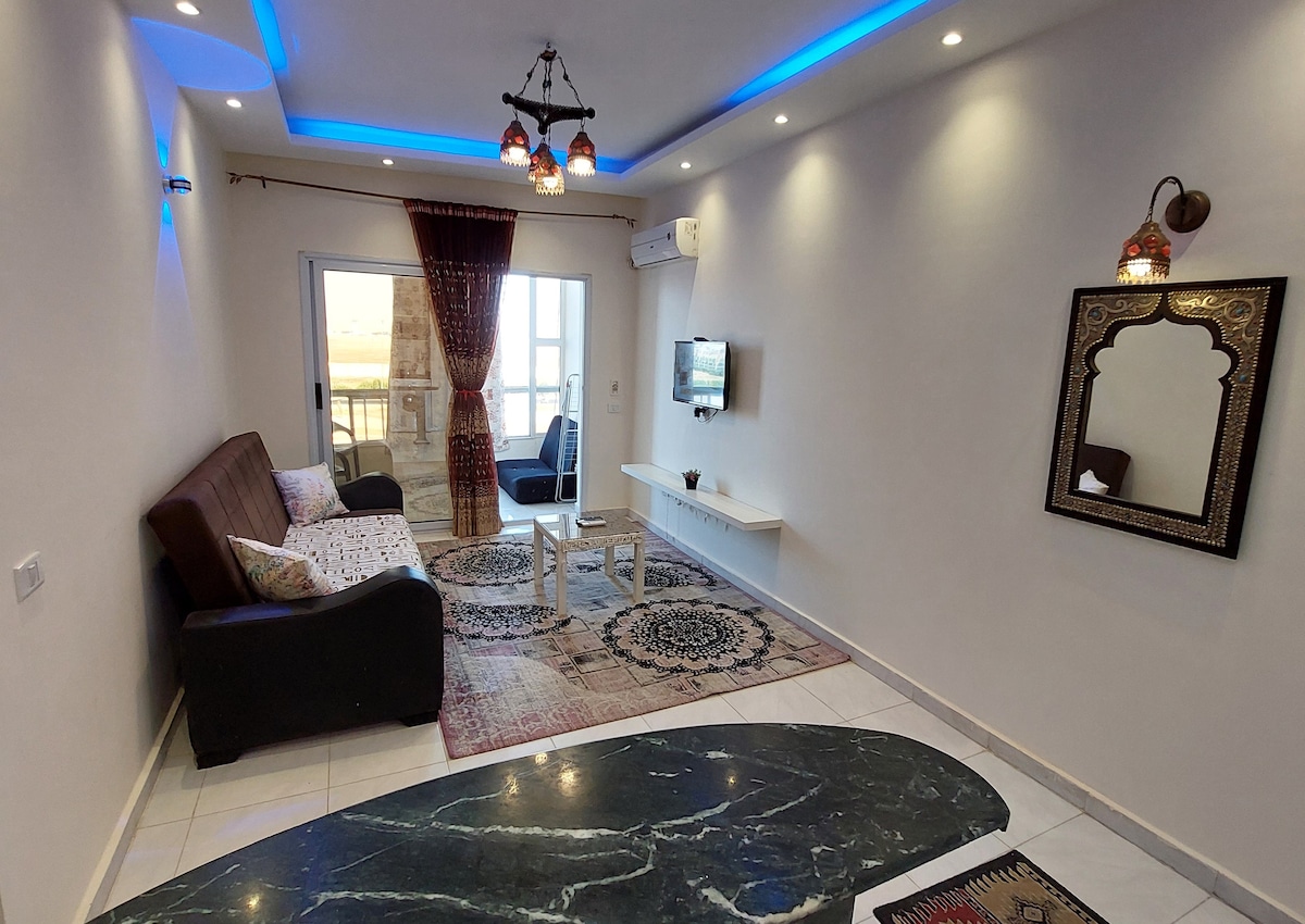 A bright living area is presented with two comfortable seating options, including a dark sofa and an armchair. A television is mounted on the wall opposite a minimalist white coffee table. Natural light enters through a sliding glass door, complemented by decorative light fixtures.