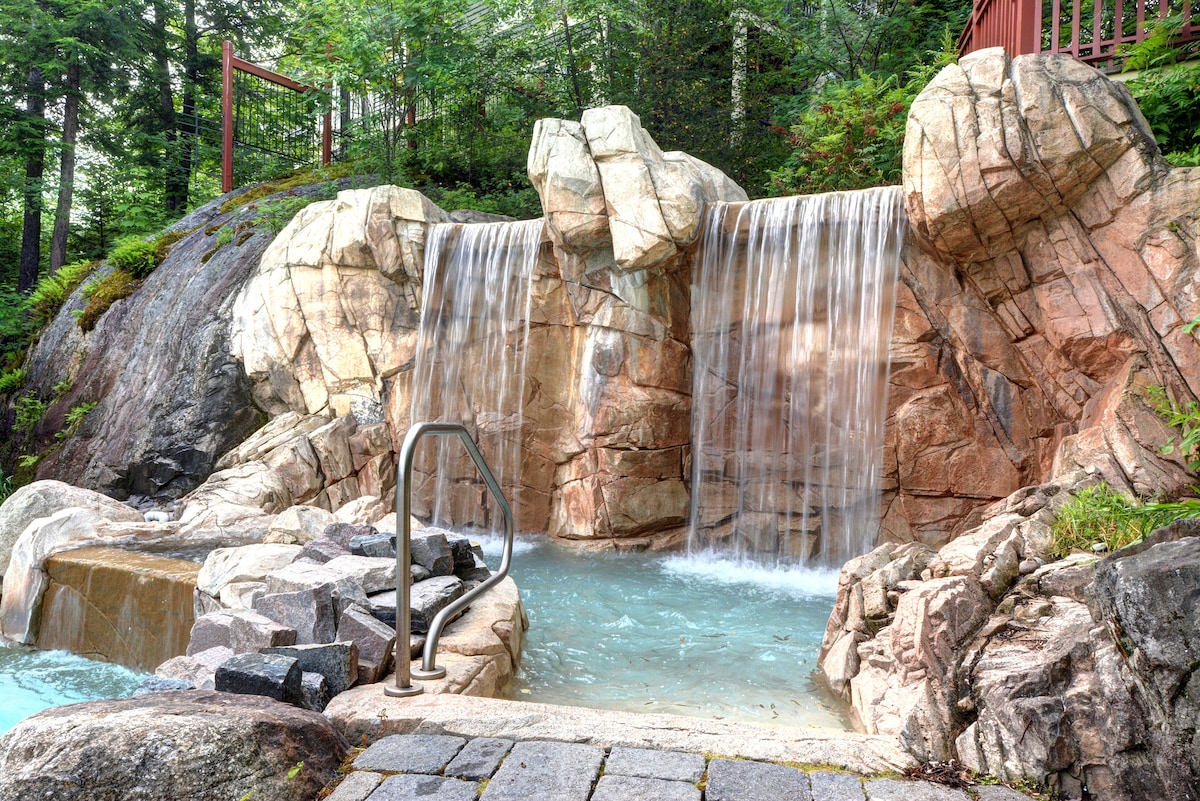 A natural stone waterfall cascades into a serene hot tub, surrounded by lush greenery. Smooth rocks and a paved area enhance the tranquil setting, inviting relaxation. A sturdy railing leads to an elevated deck in the background, blending seamlessly with the natural landscape.