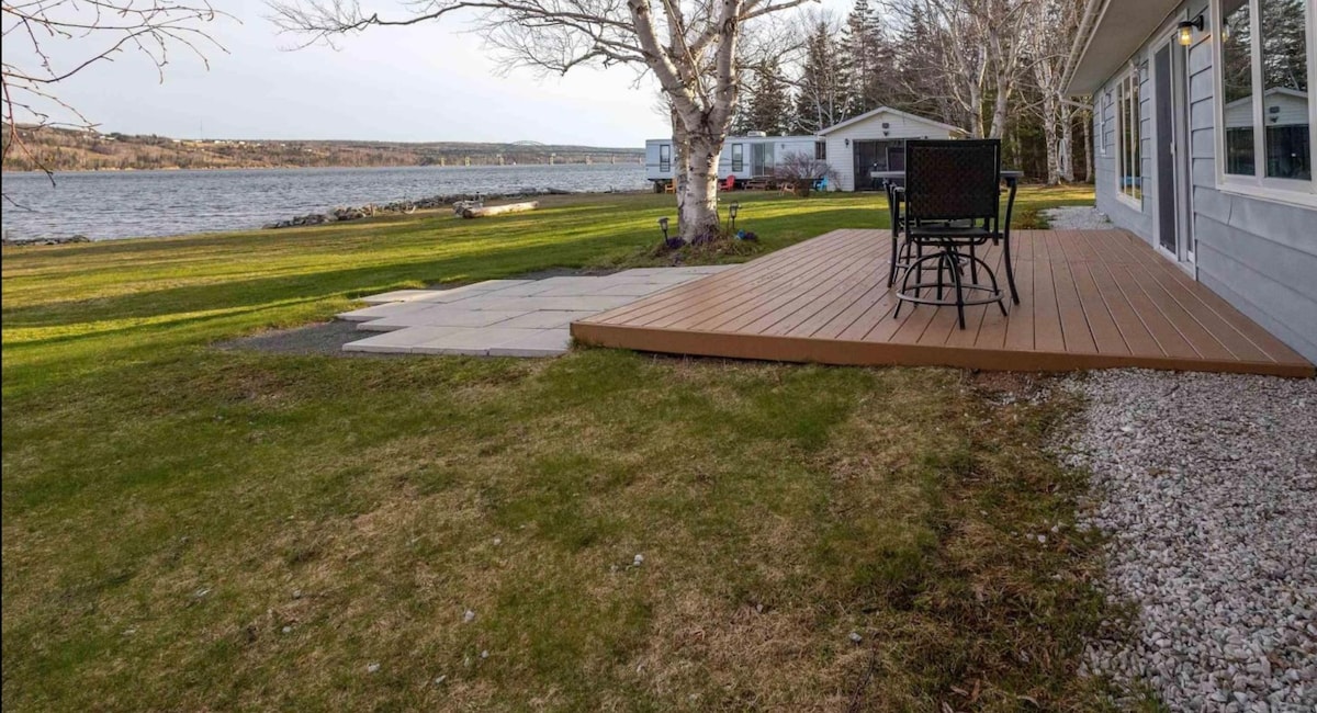 A spacious deck is positioned overlooking the Brads’Or Lakes, featuring a table with four chairs. The lawn extends toward the waterfront, bordered by trees. Gravel pathways lead to a patio area, enhancing the natural landscape.