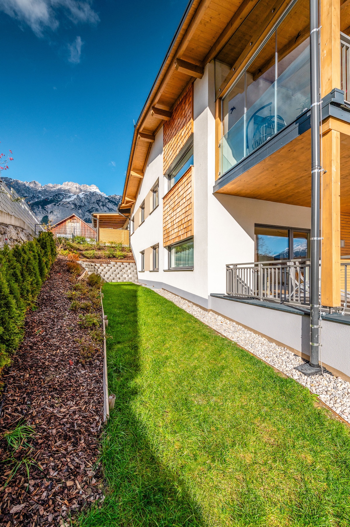 A modern apartment building is set against a backdrop of mountains. The exterior features wooden accents and large windows. A manicured lawn with gravel paths and neatly trimmed shrubs enhances the outdoor space. A balcony extends from the upper level, providing an outdoor seating area.