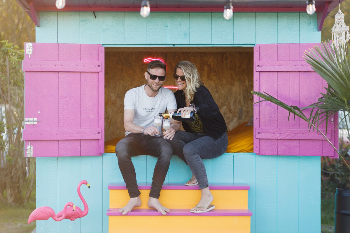 A vibrant, colorful hut features open windows framed with pink shutters, providing a welcoming atmosphere. A couple enjoys drinks on the steps, with a tropical ambiance enhanced by a pink flamingo decoration nearby. The interior is visible, showcasing a spacious bed.
