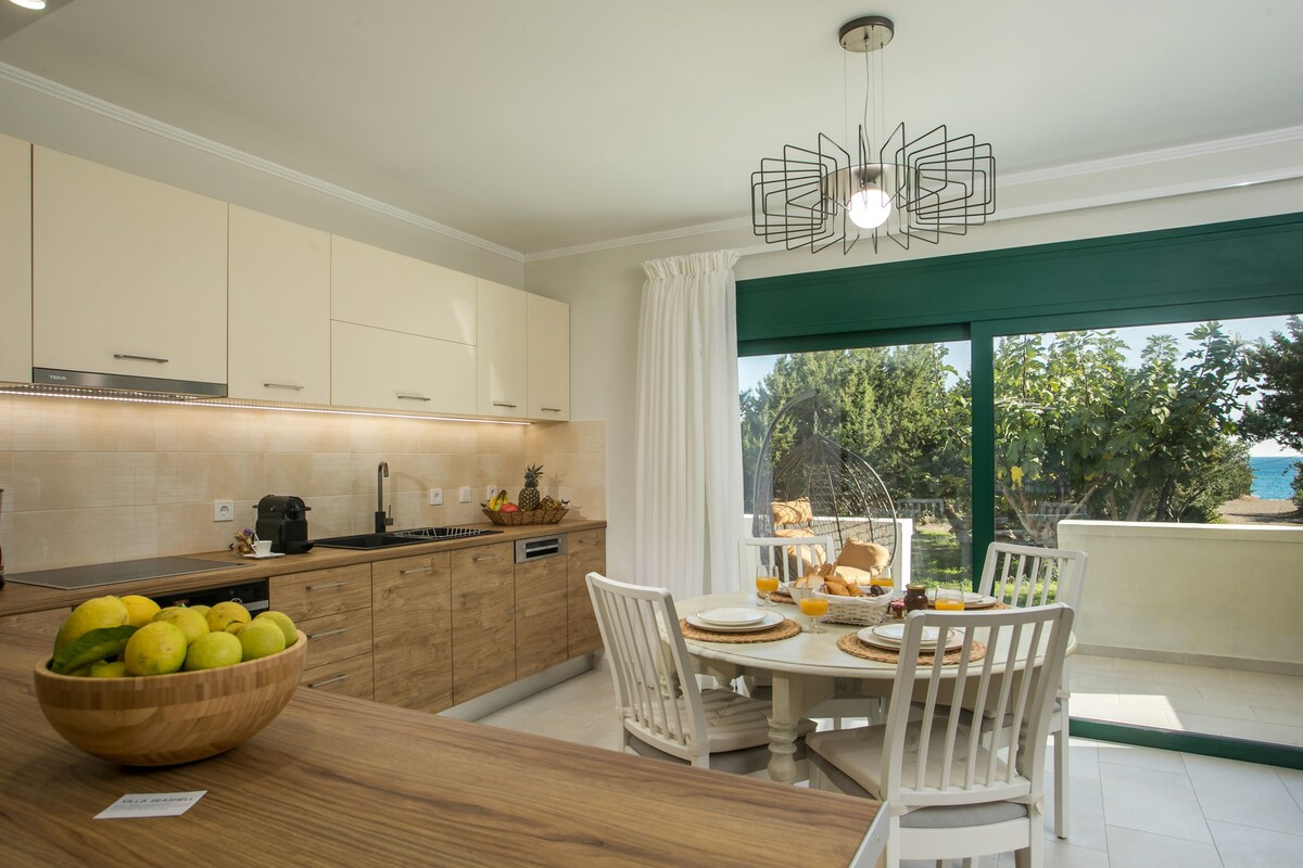 A modern kitchen area features wood cabinetry, a round dining table with white chairs, and a bowl of fresh fruits. Large windows allow natural light to fill the space, while a light fixture hangs above the table, enhancing the inviting atmosphere.