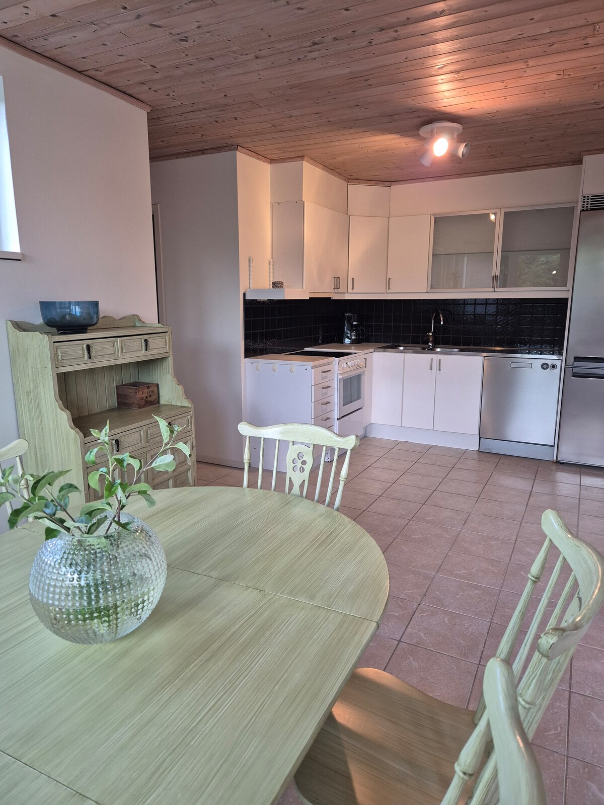 A clean and functional kitchen is presented with light cabinetry and black tiled backsplash. Essential appliances are included, along with a spacious dining table featuring a decorative vase with greenery. The wooden ceiling adds warmth to the overall space.