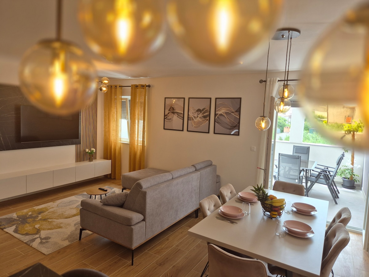 A modern living and dining area features a light-colored couch and a dining table set for six. Below hanging pendant lights, a stylish rug adds warmth to the space. Large windows provide natural light and reveal glimpses of the outdoor terrace.