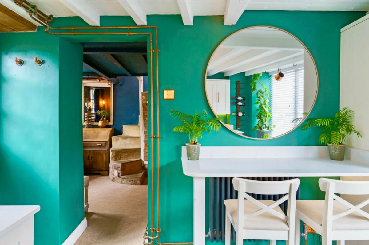 A vibrant green wall features a large round mirror reflecting the space. A small white bar with two chairs is positioned against the wall, complemented by potted plants. The background reveals a staircase and additional rooms, highlighting the cozy layout.